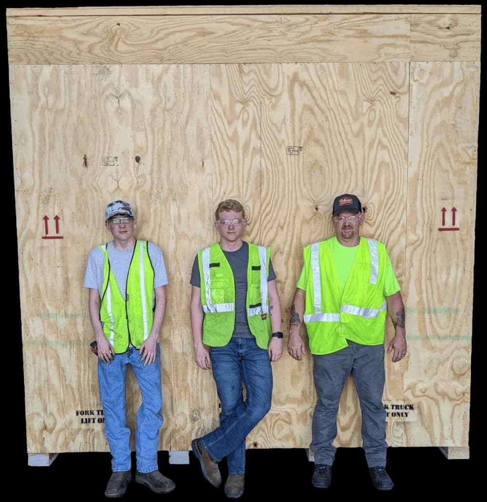 Three people in safety vests standing in front of a large wooden crate.
