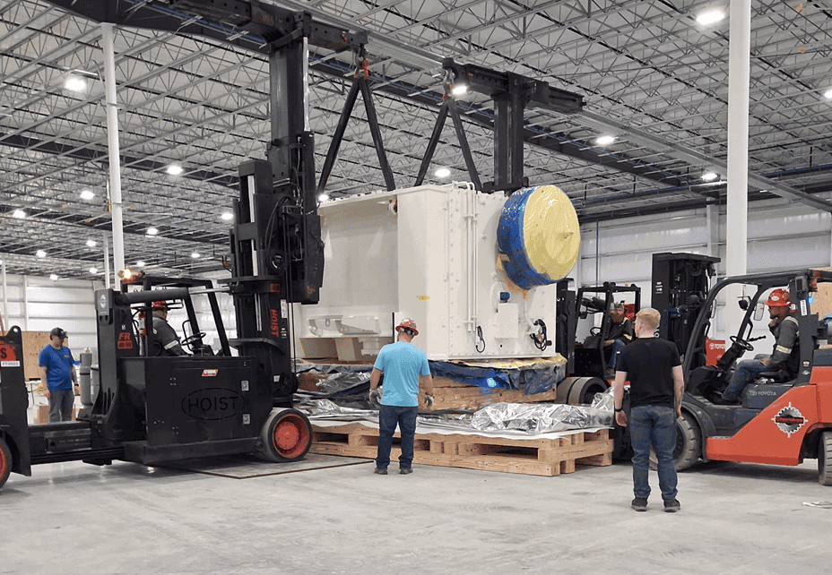 Forklifts positioning a large machine over a wood shipping base with people nearby